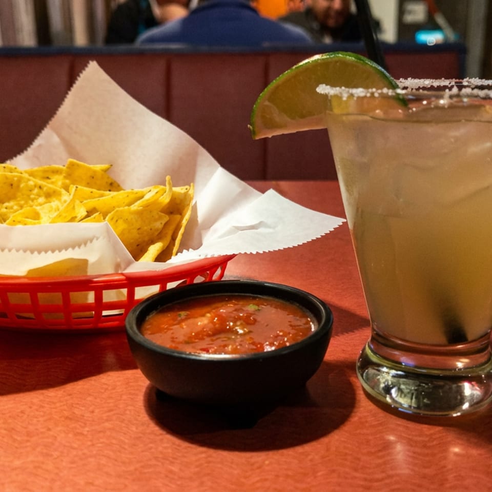 Basket of tortilla chips with salsa, a salted lime margarita, and a small bowl of dipping sauce on a restaurant table.