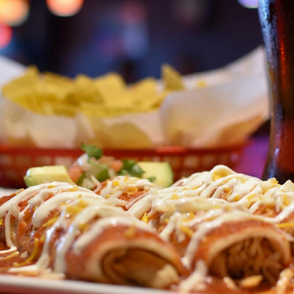 Crispy enchiladas topped with creamy sauce and cheese, served with rice, salad, fries, and a bottled soda.