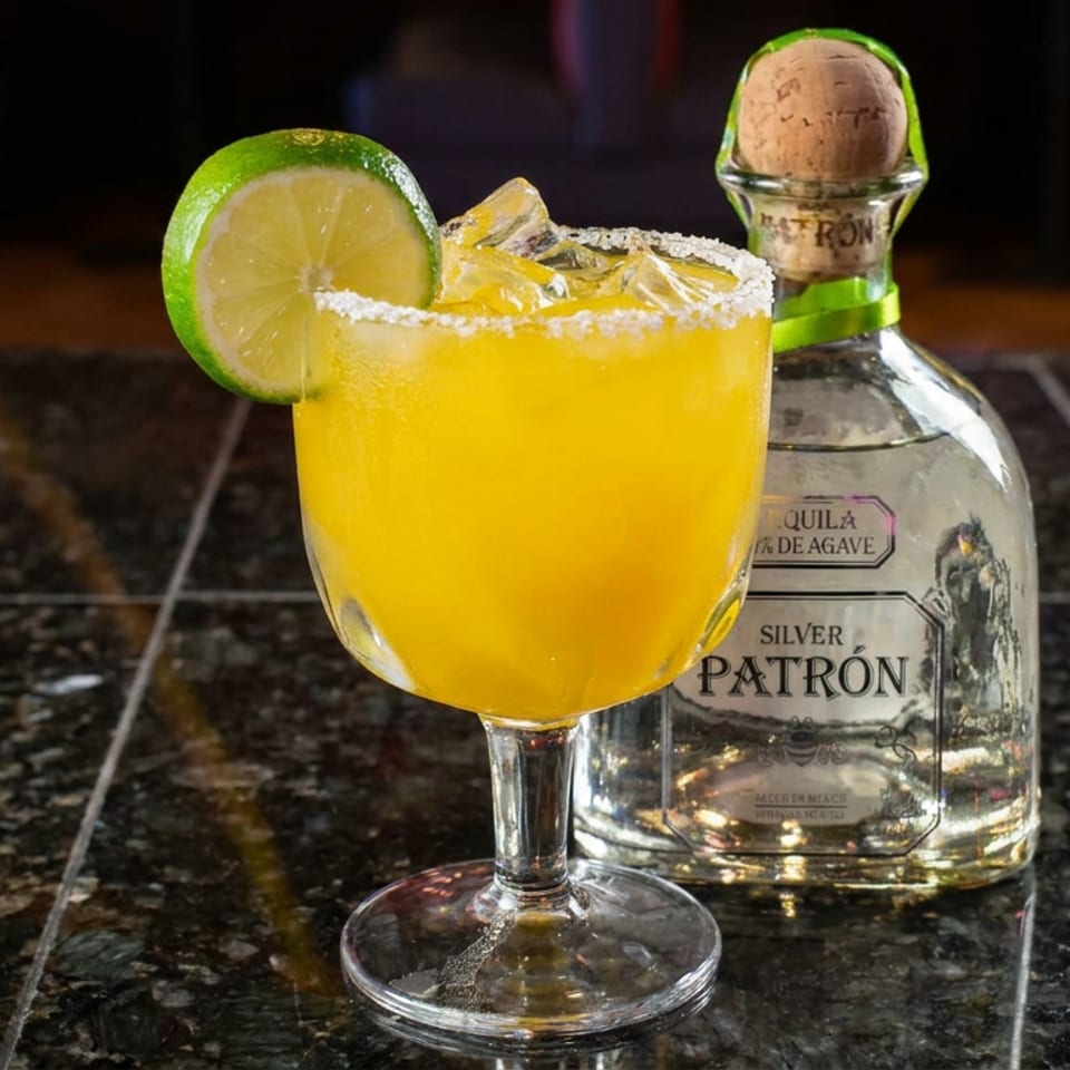 Bright yellow margarita with a salted rim and lime garnish beside a Patrón Silver tequila bottle on a restaurant bar.