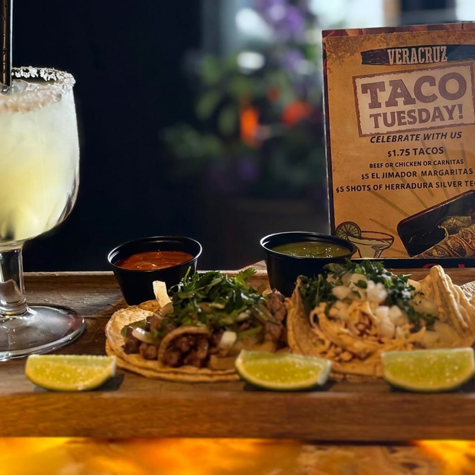 Tacos with lime wedges, dipping sauces, and a salted citrus margarita beside a Taco Tuesday promotion on a wooden board.