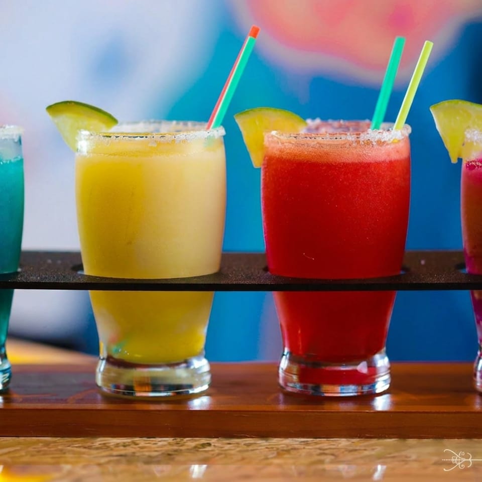 Four colorful cocktails with lime wedges and straws arranged on a wooden bar shelf, labeled Veracruz.