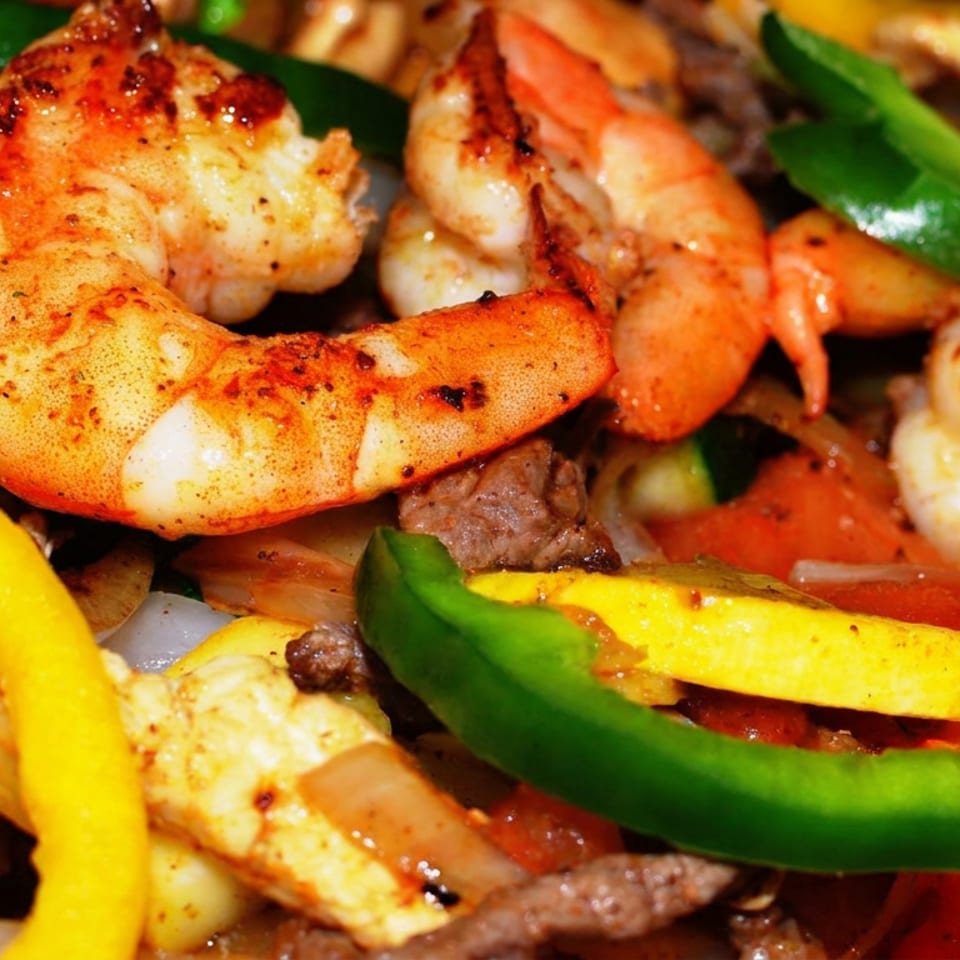 Close-up of grilled shrimp, chicken, beef, onions, and colorful bell peppers in a sizzling mixed grill dish.